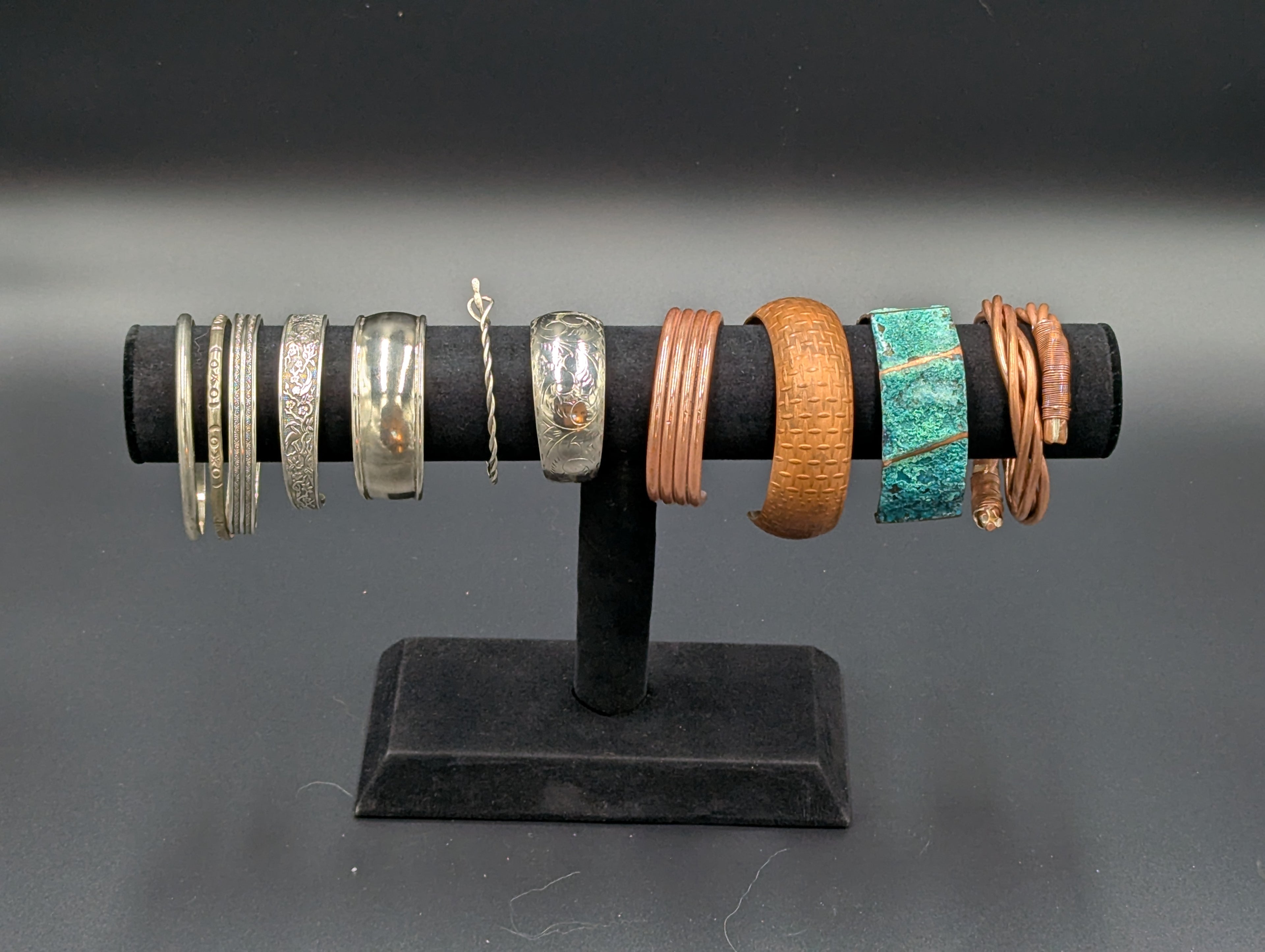 A bracelet display against a black backdrop. There are 7 silver and 4 copper bracelets on the display. They range from simple cuffs to intricate etched bracelets to treated surfaces with patina and even wire work.
