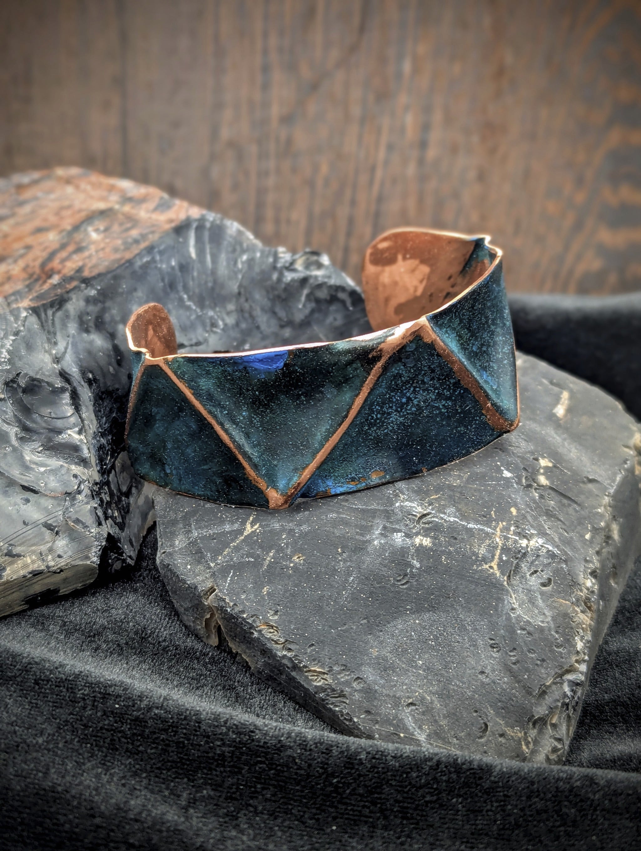Folded copper cuff with blue patina. The raised folded lines are polished copper. The cuff is sitting on black stones with a dark wood background.