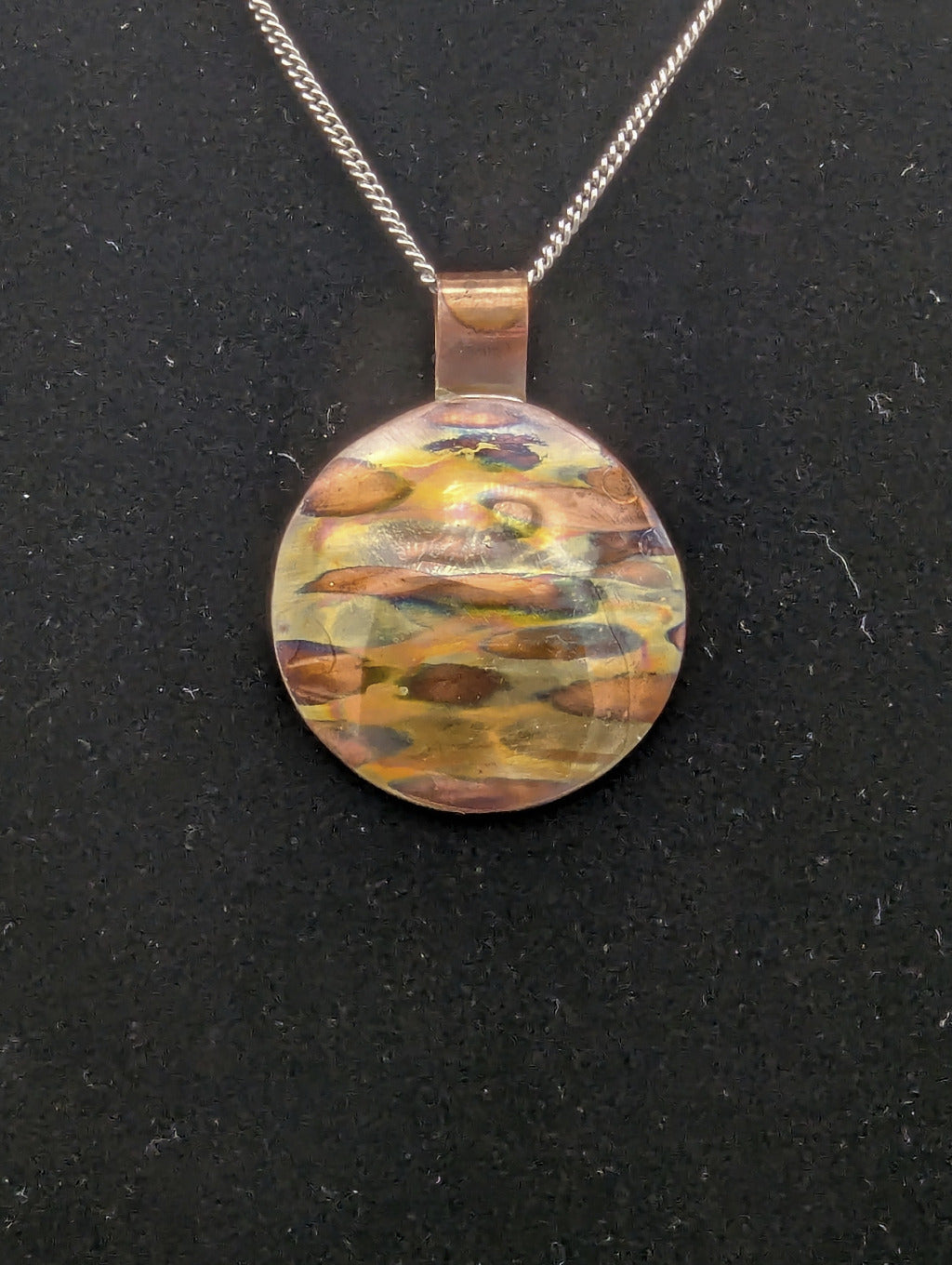 Round multicolored pendant on a chain against a black background. The pendant is copper with horizontal bands of color produced by flame painting the copper.