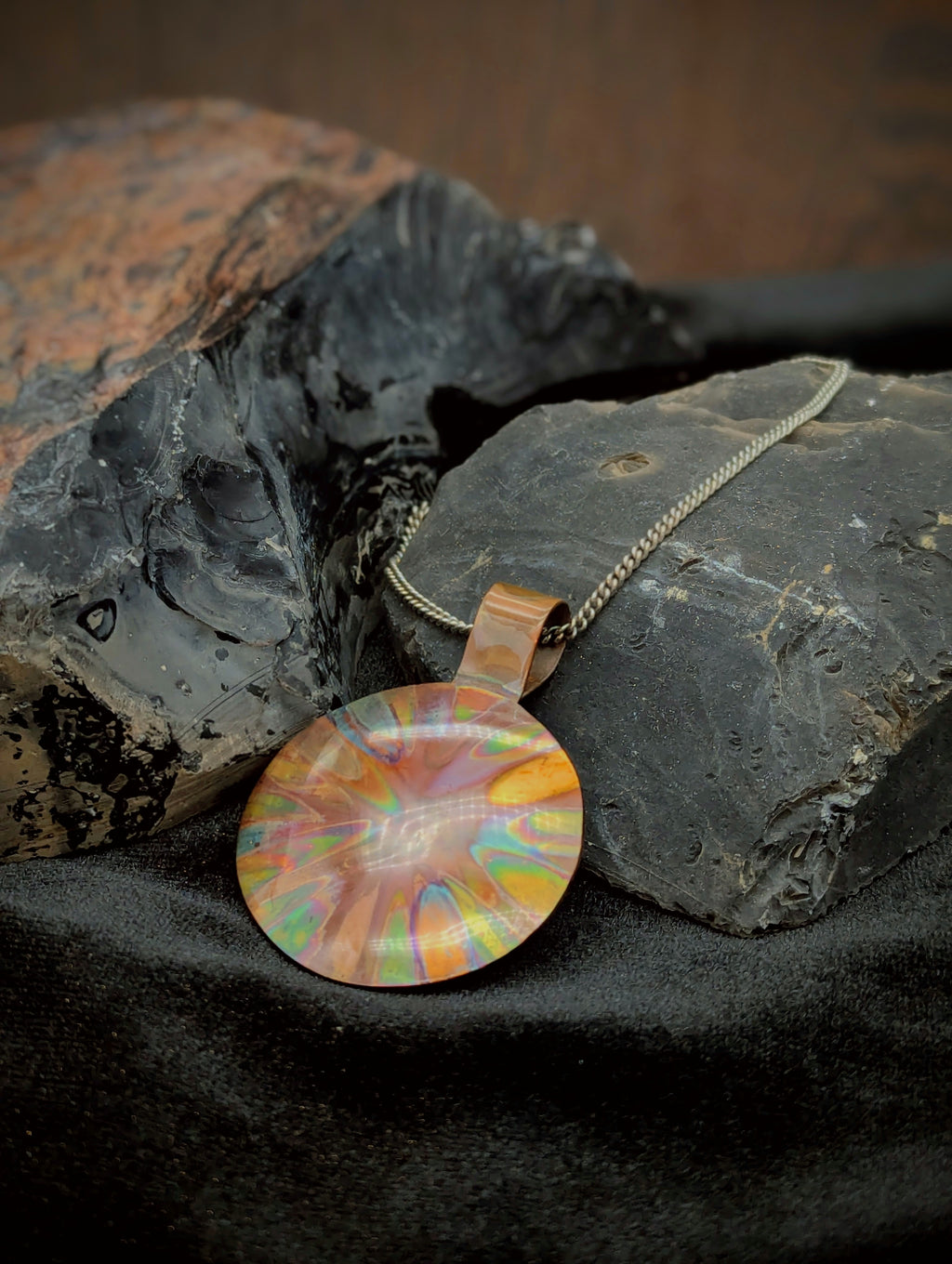 Multicolored flame painted copper pendant on a chain lying on a dark stone surface