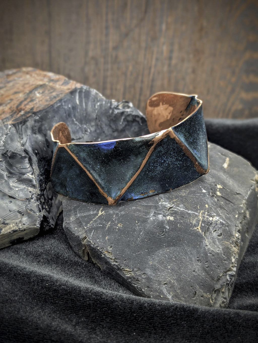 Folded copper cuff with blue patina. The raised folded lines are polished copper. The cuff is sitting on black stones with a dark wood background.