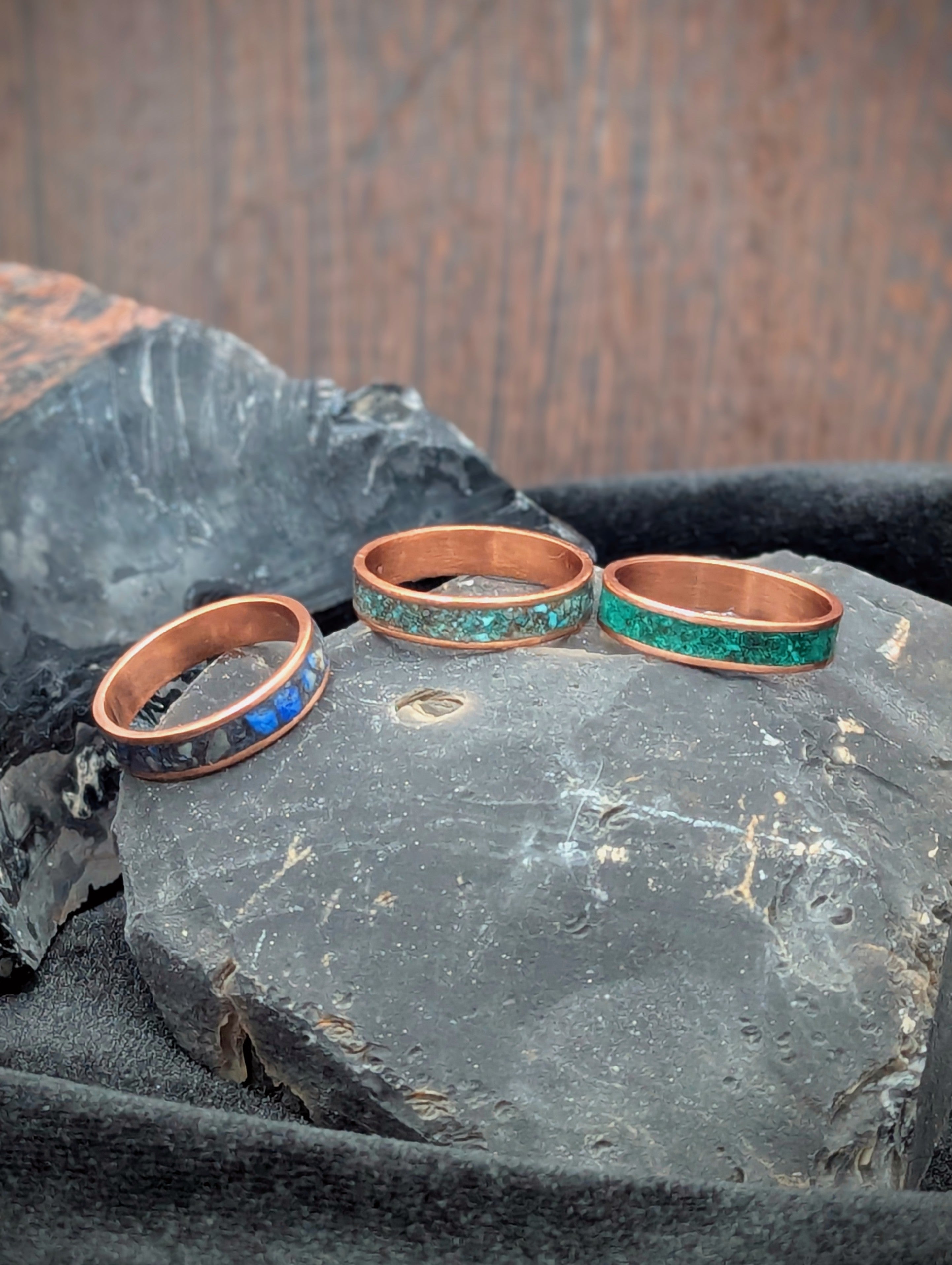 Three copper rings featuring different inlay material resting on dark stones. From left to right the inlay material is lapis lazuli, turquoise, and malachite.