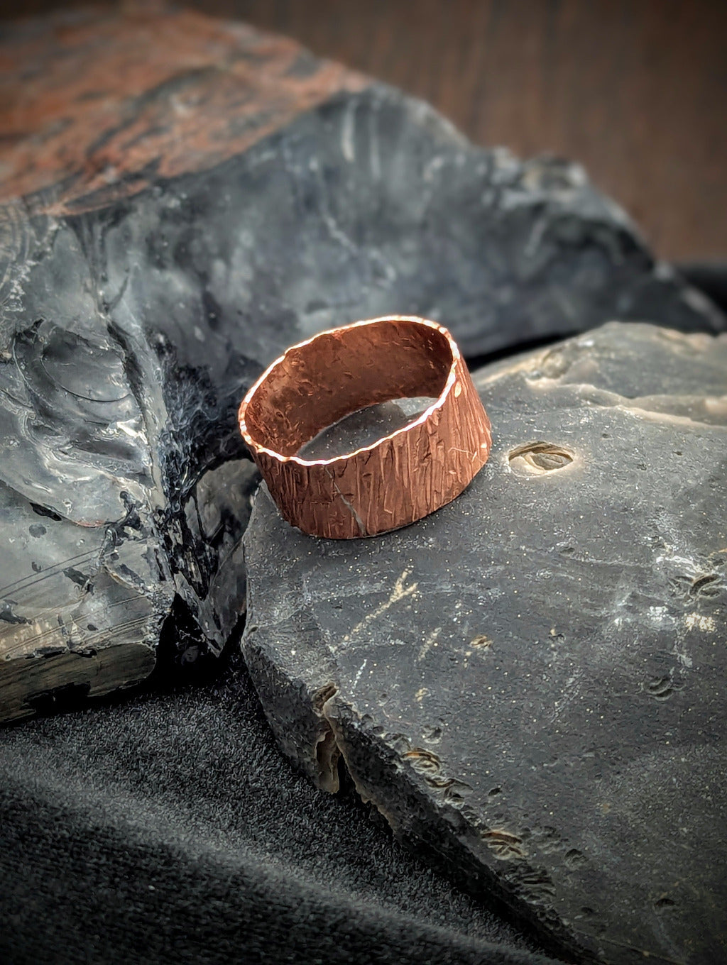 Copper ring on a textured black stone surface. The copper ring features a rich hammered texture consisting of slanted lines.
