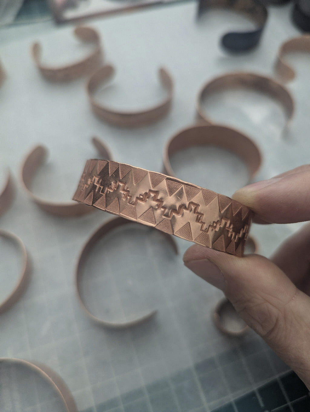 Copper bracelet with stamped geometric patterns held by a hand on a white surface with more copper bracelets.