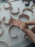 Copper bracelet with stamped geometric patterns held by a hand on a white surface with more copper bracelets.
