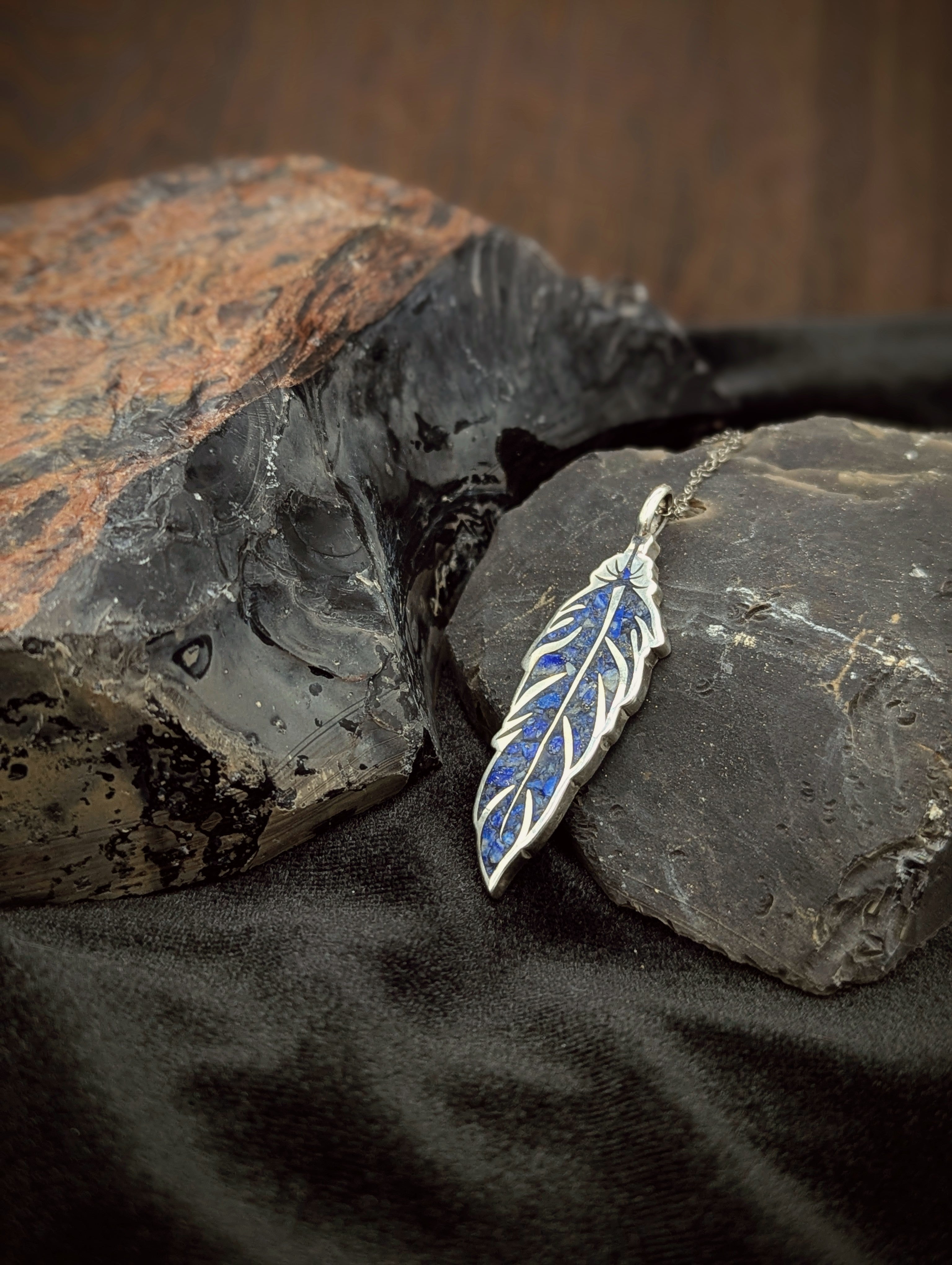 Silver feather pendant with blue stone crushed inlay on a dark stone surface