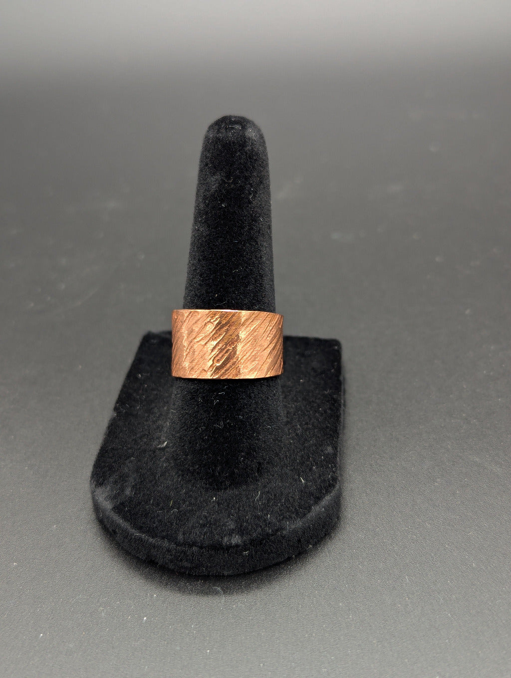 Copper ring on a black felt stand against a gray or black backdrop. The copper ring features a hammered texture of slanted lines.