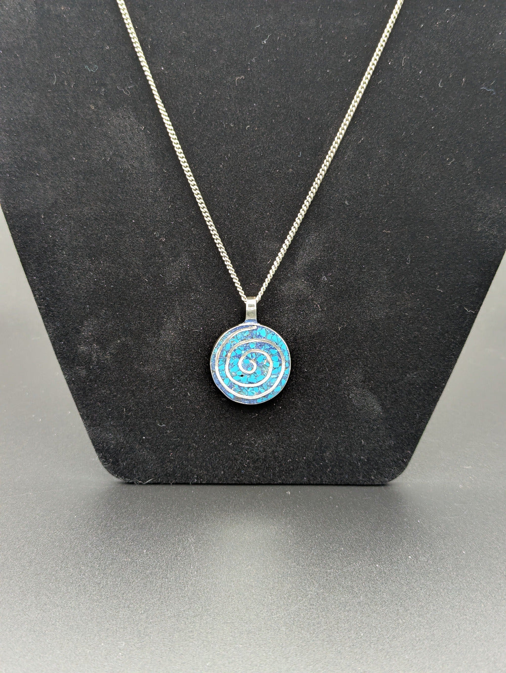 Necklace with a blue and silver spiral pendant on a black stand. The blue is from turqoise inlay.