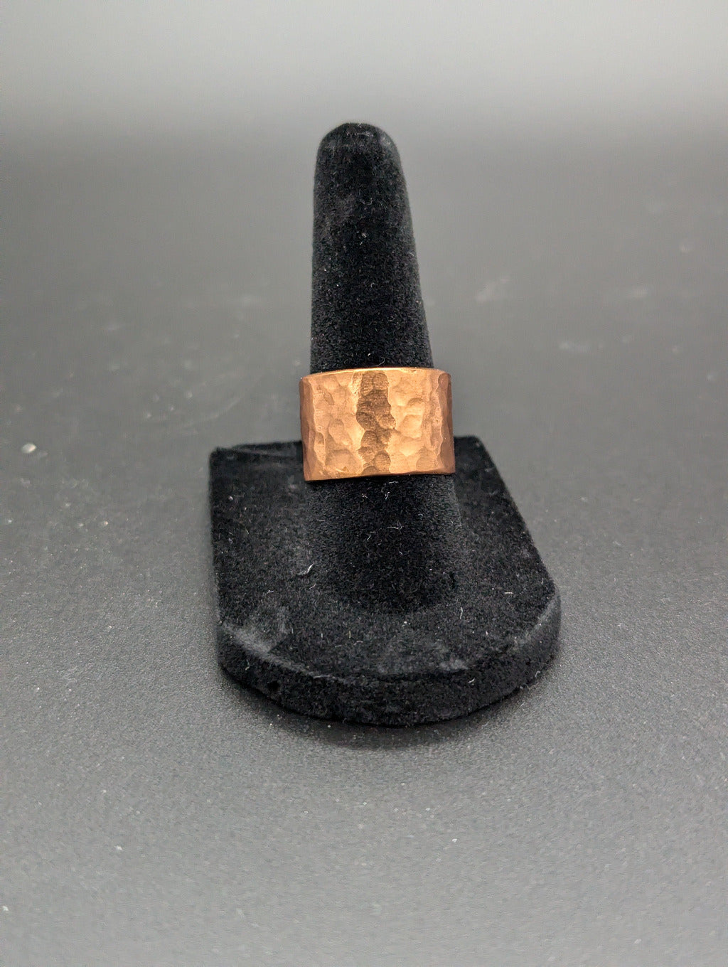 Copper ring on a black stand against a gray background. The copper ring features a striking hammered pattern resembling a mule mug.
