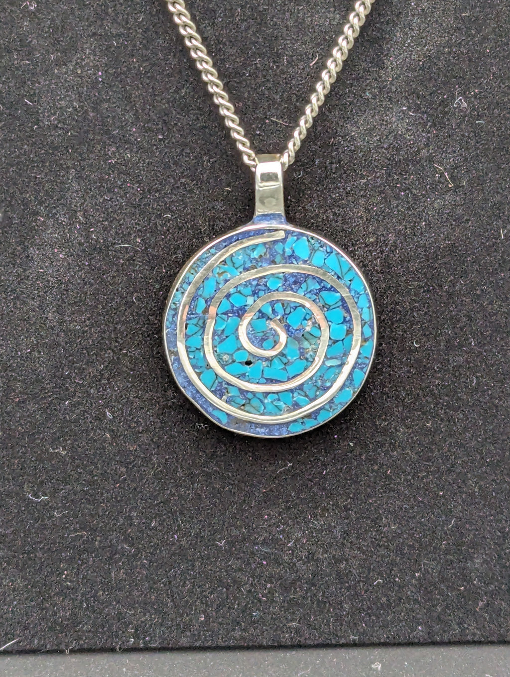 Sterling silver circular pendant on a black felt background. The pendant includes a silver spiral and is filled with turquoise and various dark blue inlay.