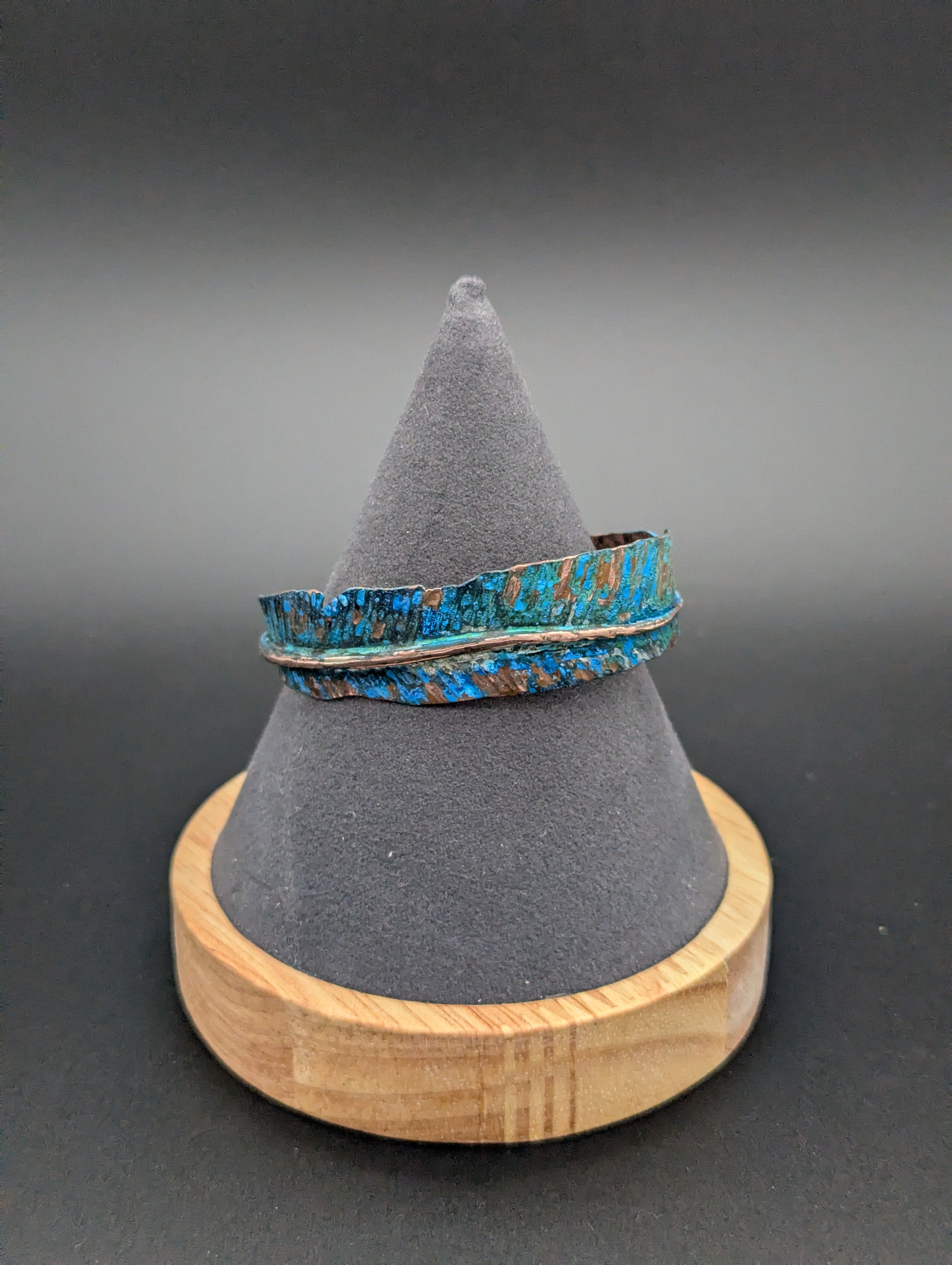 Handmade copper cuff shaped like a feather. Blue patina covers some of the feather in between the groves of the hammered feather texture. The cuff is sitting on a gray felt cone.